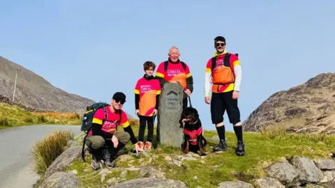 James Bavington The Bavington family including 8-year-old boy Harry, stand around a stone maker on Snowdon as they walk up the mountain. They are wearing bright pink and orange t-shirts which say Breast Cancer Now.
