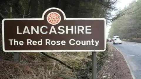 A Lancashire road sign. It is brown with white letters saying Lancashire, The Red Rose County. There is a road to the left of it with cars on it.