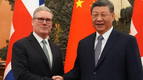 PA Media Sir Keir Starmer, who has grey hair and glasses, is wearing a dark suit and green tie, shaking hands with President Xi, who has black and grey hair which is neatly styled. They are standing in front of a large Union Jack and Chinese flag.
