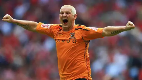 Getty Images Windass memiliki rambut pirang pendek yang diputihkan dan mengalir dengan kedua lengan di luar di atas lapangan di Wembley. Dia mengenakan kemeja jeruk Hull City.