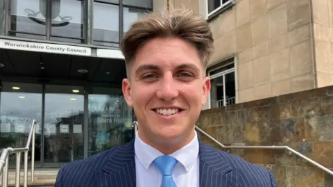 George Finch stands outside the headquarters of Warwickshire County Council. He wears a pin stripe blue suit jacket, white shirt and blue tie.