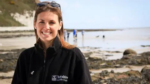 A woman with long brown hair and black sunglasses resting of her head is smiling into the camera. She is wearing a black Yorkshire Wildlife Trust fleece. The background is blurred but you can see rocks on a beach next to cliffs and a group of people near the water.