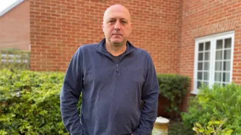 LDRS Colin Phillips stands outside a modern brick-built house, with a hedge and shrubs behind him. There is a white window in the side of the house. He is bald and wearing a navy blue open-collared jersey top.