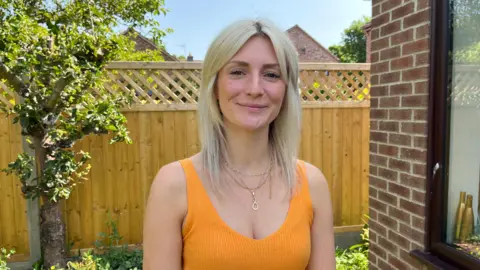 BBC/Shaun Whitmore Emma-Jayne Herbert is smiling at the camera, wearing an orange top standing in the garden.