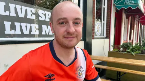 BBC/Nicola Haseler Jack O'Shea with very short brown hair and beard wearing a Luton shirt