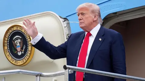 Getty Images Donald Trump, wearing a dark suit, white shirt and red tie, waves as he emerges from the presidential plane Air Force One. 