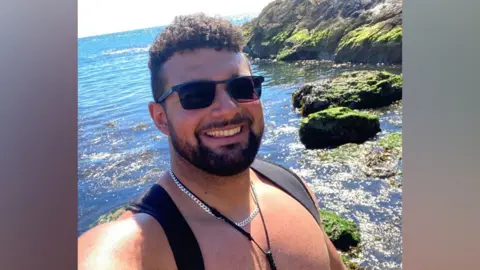 A man with short curly brown hair and a short dark beard smiles as he poses for a selfie in front of the sea and a rocky cliff. The sun is shining and he is topless wearing sunglasses, a silver chain and backpack straps.