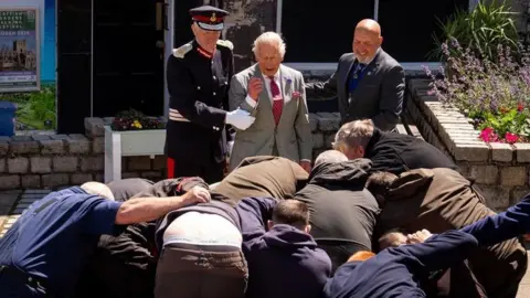 King Charles has arm held by uniformed officer as men scramble in a scrum in front of him