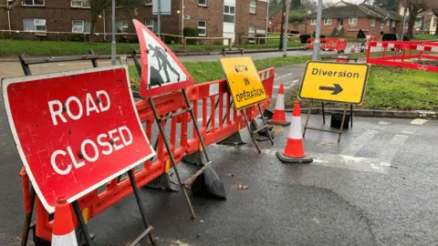 Henry Godfrey-Evans/BBC Road closed signs, diversion signs and orange barriers blocking the road. There are flats in the background.