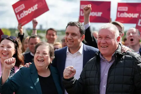 Getty Images Dame Jackie Baillie, Anas Sarwar and Davy Russell stand in a row, smiling and appearing to punch the air in joy at their election win. The first two are in business wear, Russell is in a puffer jacket and a checked shirt. There are many happy people behind with red Labour flags.
