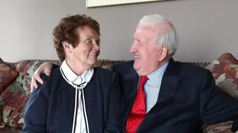 Sir Kenneth Bloomfield has grey hair and is wearing a dark suit with a red tie. He is sitting on a red and brown sofa and is smiling at his wife who has short dark hair and is wearing a white blouse with a navy cardigan and is smiling back at him. They are both in their 80s.