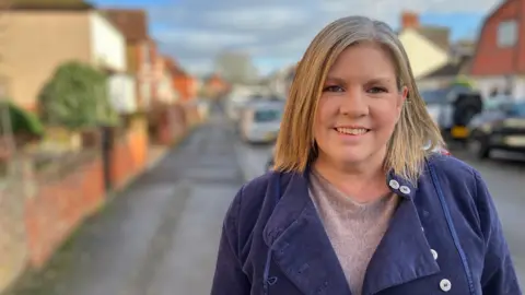 Melissa Harris smiles for the camera, standing in her street. She has straight, fair, shoulder-length hair and wears an indigo coat over a beige jumper.