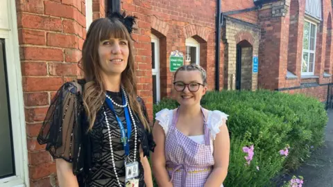 James Grant/BBC A woman in a black lacy dress stands next to a woman in a white dress with a squared apron. They are smiling and are in front of a brick building. 
