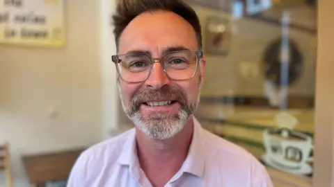 A man in a white shirt and glasses with a grey beard and brown hair.