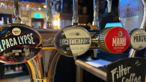 The photo has four beer handpumps in a pub. In the middle a pump has the Hot Fuzz logo with a black and white background. Behind the bar there are wine glasses hanging upside down on a rack, wine bottles on a bar and a mini fridge with cans inside. 