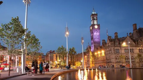 Computer enhanced image of the city centre including the clock tower lit up purple and lampposts reflecting light off the water.