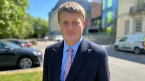 Richard Clewer faces the camera in a suit, standing outside a sunny County Hall