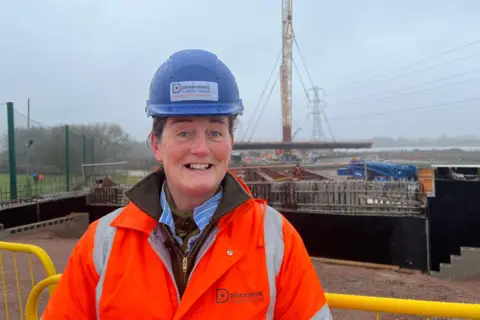 A woman on a construction site in a orange hi-vis jacket and blue hard hat. The words DERBYSHIRE COUNTY COUNCIL are on the jacket and hat.