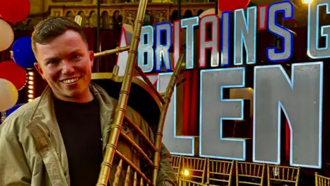 Sonny Green smiling at the camera holding a golden chair over his shoulder with the Britain's Got Talent logo and words in the background. He is wearing a khaki or golden-coloured jacket. He has short brown hair.