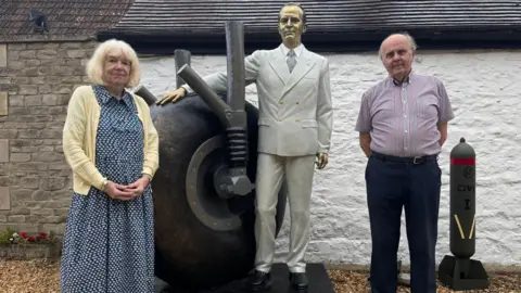 BBC Martin Robins and Caroline Flippants stand next to a bronze statue of Sir George Dowty which depicts him dressed in a light-coloured suit with one hand resting on an aeroplane wheel