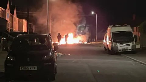 PA Media A fire in a dark street with people in front of it