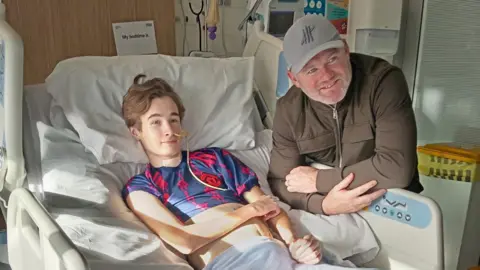 Julie Clewes A teenage boy wearing a red and grey Manchester United shirt is lay down in a hospital bed. Wayne Rooney leans over the side of the bed smiling while wearing a grey cap and a khaki green jacket.