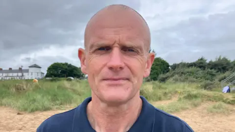 Keiron Tourish A man with a shaved head is looking at the camera with a netural expression on his face. His head and shoulders are visible. He is wearing a blue top, what appears to be a polo shirt. He is standing outside on a beach, in the background sand, grass and bushes can be seen. 