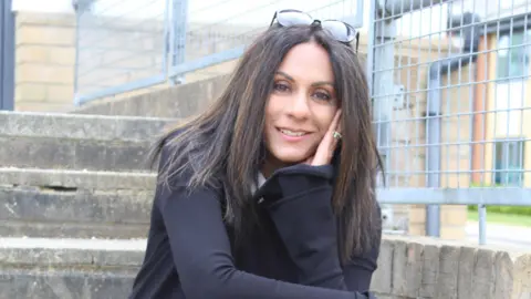 Kathryn Croft Kathryn Croft, a woman with long dark hair, wears a black jacket and poses on a set of concrete steps in front of a metal railing, her glasses pushed up on the top of her head, as she smiles into the camera.