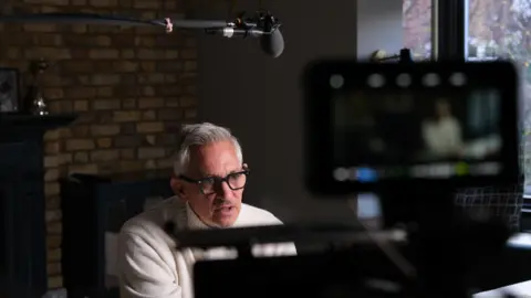 Sylver Entertainment/ITV A man with grey hair, glasses and white turtleneck jumper sits in a chair in front of cameras. There is a microphone above him.