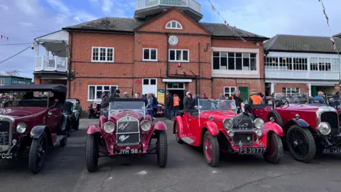 Brooklands Museum Four red vintage motors are parked in front of Brooklands Museum - a red brick building with a white viewing platform on the side.