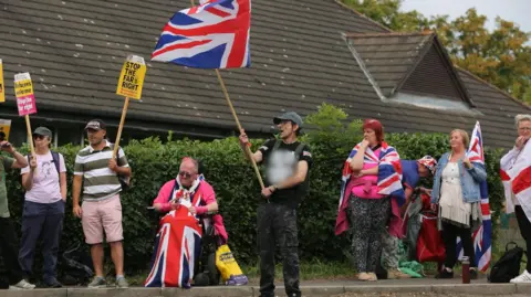 Protestors waving flags and counter protesters with signs saying 'Stop the far right' gather outside a hedge with a hotel behind it