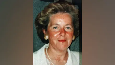 A photo of the late Elizabeth Rush who had brown hair, green eyes, and was wearing a white shirt with a pearl necklace and gold earrings.