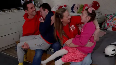 A father sits on the sofa with his son on his knee and a mother sits with her daughter on her knee. They are smiling and rocking. The boy, with dark brown hair, has grey jogging bottoms and a navy top and the girl, with dark hair in a pink bow, wears a pink hoodie and pink skirt and white tights. Behind them is a row of bunny teddy bears. The parents are wearing red hoodies with Ollie's Army written on them. The dad has dark hair and the mum has blonde hair. 