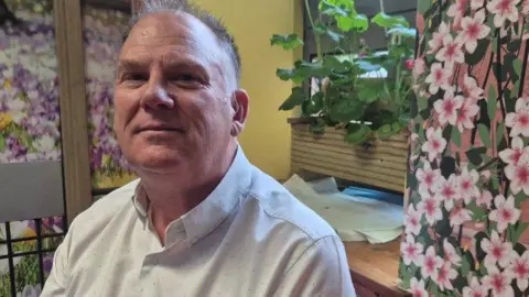 Douglas is smiling at the camera and is wearing a white shirt which is open at the collar and has short grey hair. He is sat in front of pink flowery wallpaper, a green plant and a wooden bench.