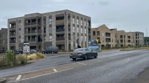 Tom Jackson/BBC Several flats which are part of a four-storey bricked building. There is a road which runs in front, and there are several cars on the road.