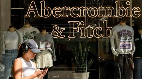 Getty Images, una mujer joven con una gorra de béisbol de los Yankees de la Marina de Nueva York y vence a los auriculares, con una camiseta sin mangas blanca, pasa junto a una tienda Abercrombie & Fitch en la ciudad de Nueva York. Tiene el nombre de la marca en Neon en la ventana y la pantalla presenta ropa informal como sudaderas grises con el logotipo de los New York Giants.