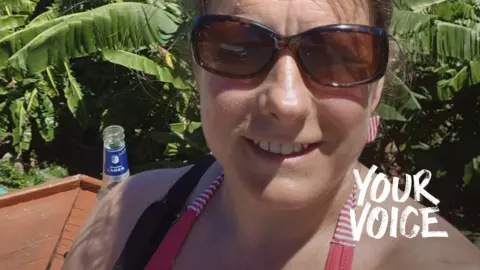 Flora Badger Flora Badger, wearing a red halter top and dark brown sunglasses, smiles taking a selfie. Behind her there is a view of the sea.