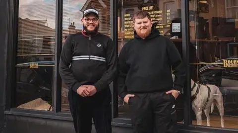 Two men standing side-by-side outside the glass front of a barber shop. The man on the left is wearing a dark zip-up jacket and a dark hat. The man on the right is wearing a black hoodie.