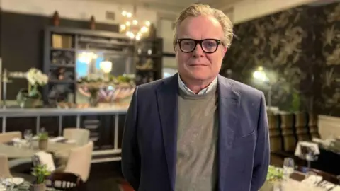 A man with slicked back blond, greying, hair and black-rimmed glasses looks straight down the camera. He's wearing a navy jacket over a grey jumper and white dress shirt. He's standing in a restaurant - behind him are tables and chairs with small green plants. 