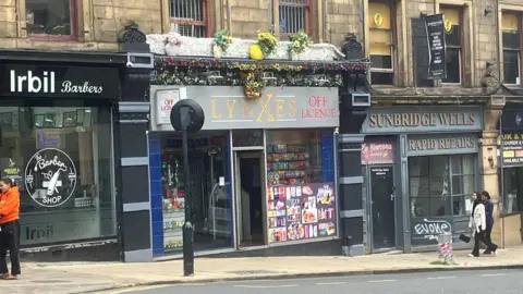 LDRS A street-level view of a row of small shops in an older stone building. In the centre is a shop with a sign reading “LYNXES OFF LICENCE” in gold lettering, with a window display filled with brightly coloured products such as greeting cards and small items. The shopfront is decorated with artificial flowers and greenery above the sign. To the left is a barbershop with signage reading “Irbil Barbers” and a circular barber logo on the window. To the right, neighbouring businesses display signs reading “SUNBRIDGE WELLS” and “RAPID REPAIRS.” A black post stands on the pavement in front of the shops, and metal barriers line the street edge. Two pedestrians walk past on the pavement, and the overall scene appears to be a typical urban high street.
Provide your feedback on BizChat