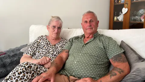 BBC/Shaun Whitmore Nicki and Andy Duffield are sitting on a sofa looking at the camera. Nicki is wearing a white patterned dress and glasses. Andy is wearing a green and white patterned shirt.