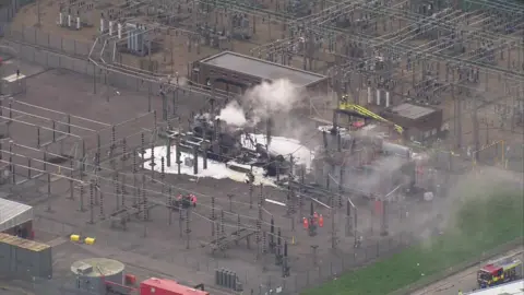 An aerial view of the substation and power lines around it in the morning when the fire died down but was still smoking, with a yellow turntable ladder extended over the scene and various staff in fluorescent red uniforms standing nearby in groups