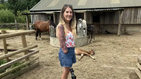 Caroline stands looking at the camera. She has long blond hair held back by sunglasses on her head. She is wearing denim shorts and a floral vest top. Caroline has a metal prosthetic leg and her arm below her elbow has been amputated. In the background are two horses and two foals, with a barn behind.