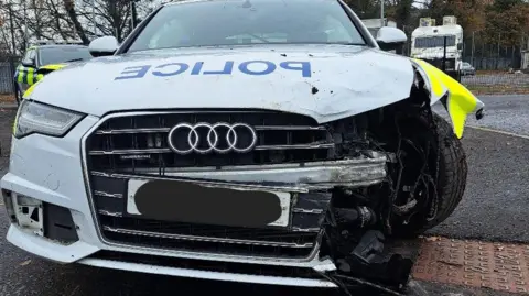 A PSNI car with damage to the front of the vehicle. The front of the car is ripped off and the is visible. 