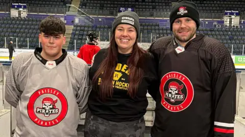 A woman with long red hair wearing a Boston Raiders jumper from Heated Rivalry stands between two men wearing Paisley Pirates shirts and ice hockey gear as all three stand in front of an ice rink.