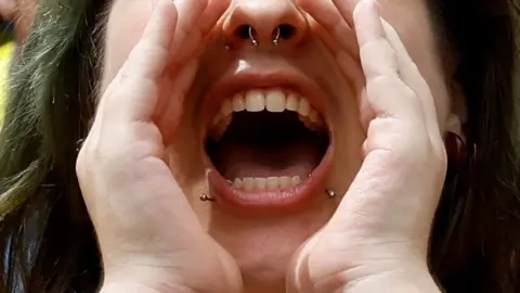Reuters A woman with nose and lip piercings cupping her mouth with her hands while shouting