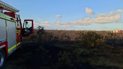 HIWFRS Fire engine to the left of the picture and another in the distance on heathland where smoke can be seen rising due to a heath fire.
