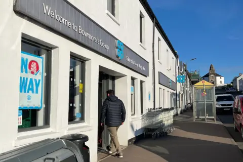 BBC The front entrance of a Co-op grocery shop in Bowmore on Islay. A man wearing a blue puffer jacket and light chinos, with his back to the camera, walks in through the doorway.