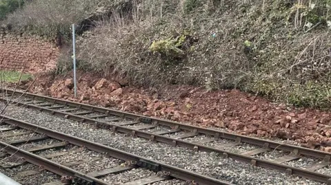 A grassy bank close to the railway line has partially collapsed with rubble resting next to the line. 