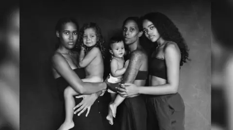 Aniya Emtage / Eye Mama Project Aniya Emtage photographed with her family, a baby, a young girl, and two older sisters in a black and white image.